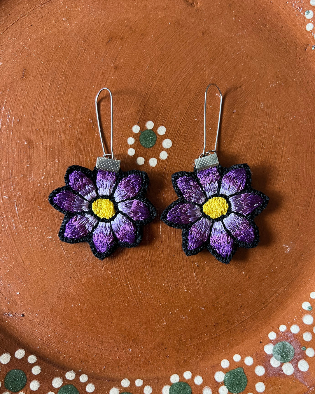 Pair of purple flower earrings on a barro plate surface with decorative elements.