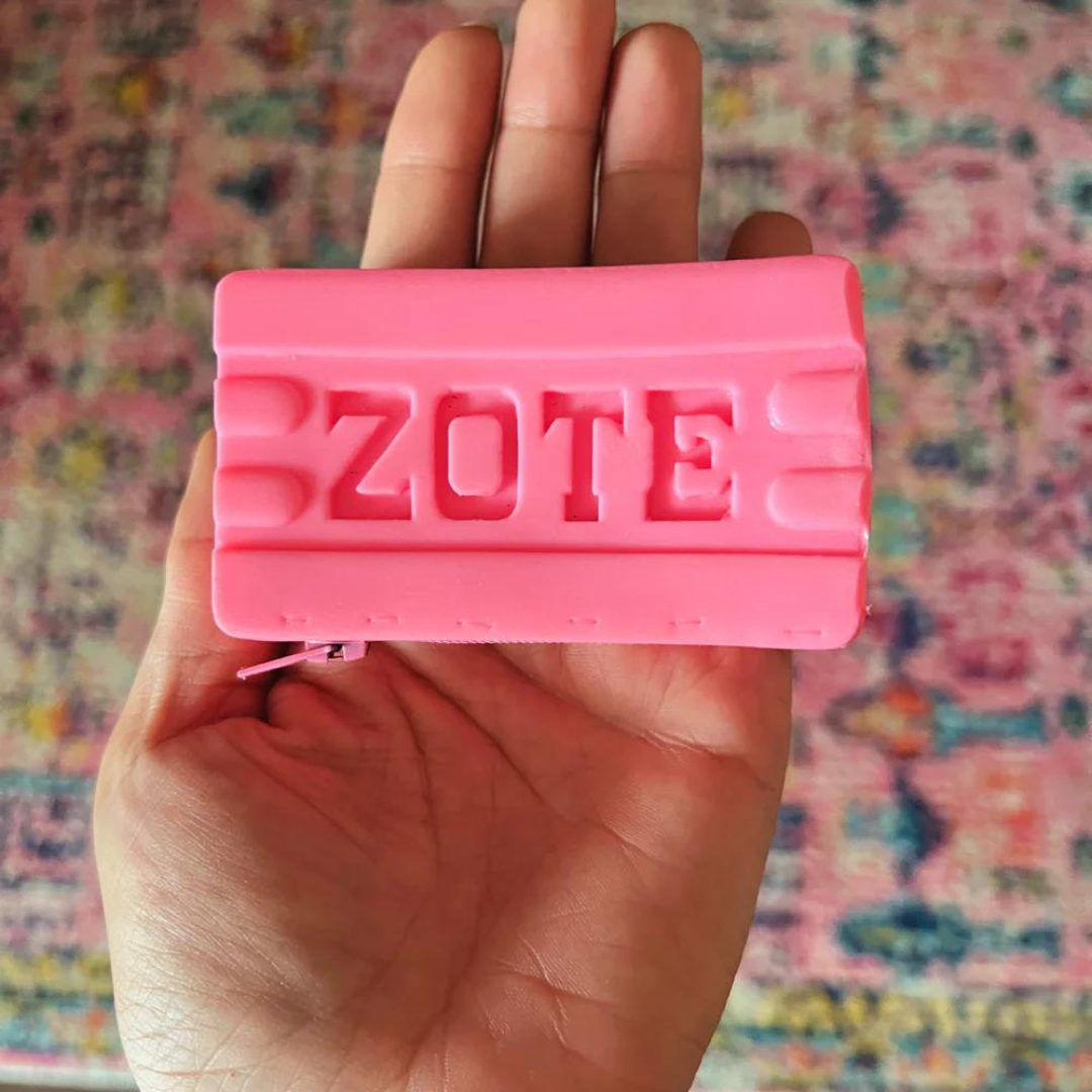 A hand holding a pink coin purse with the word 'ZOTE' embossed on it, against a patterned background.