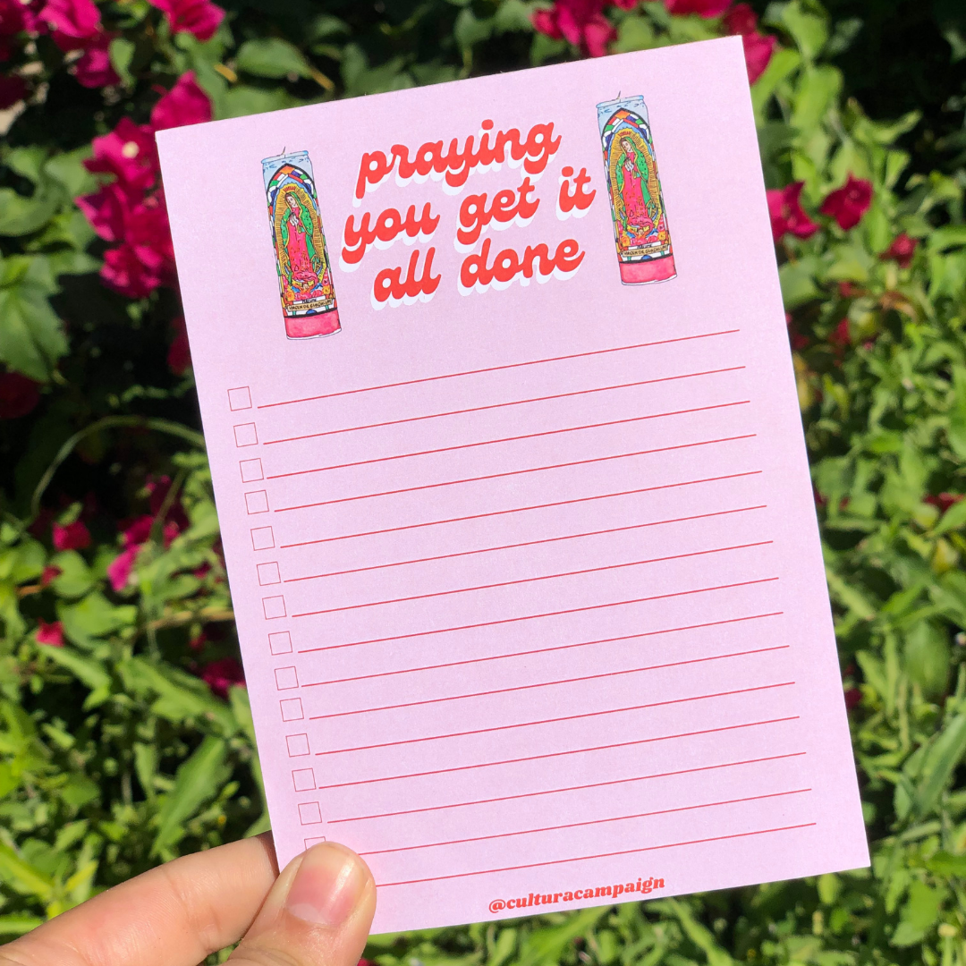 A pink notepad with a design and text on the cover, held in a hand with a background of flowers.