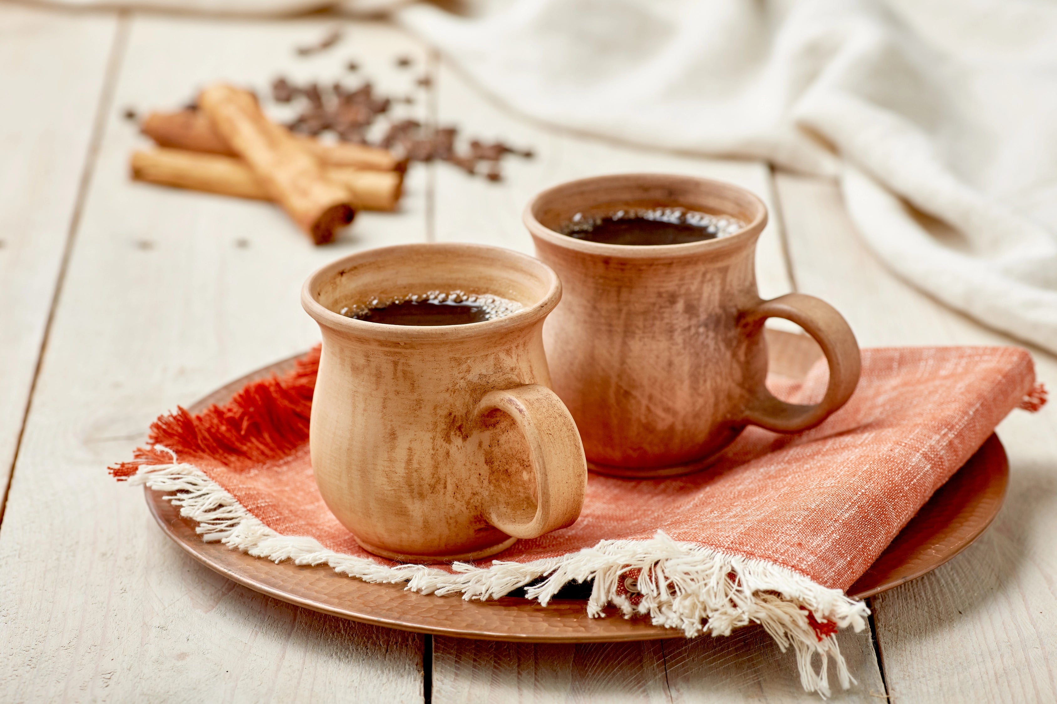 Two ceramic mugs with coffee on a wooden table with cinnamon sticks and coffee beans.