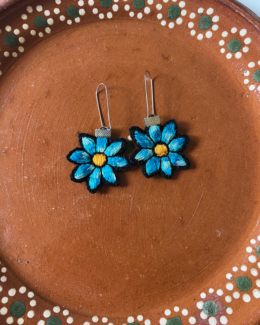 Pair of blue flower-shaped earrings on a barro ceramic plate with decorative edge.